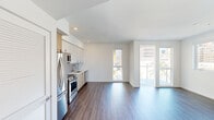 This modern urban residence is situated within a striking multi-story building featuring a bold exterior palette of yellow, gray, and blue. The single-floor layout is designed with clean lines and a bright, contemporary aesthetic throughout. The open-plan living and dining area is anchored by rich hardwood-style flooring and crisp white walls, creating a versatile and airy space. Large windows and a glass-railed balcony door flood the interior with natural light while offering expansive city skyline views, including surrounding commercial and residential towers. The kitchen is thoughtfully appointed with a seamless combination of white upper cabinetry and warm wood-toned lower cabinets, complemented by a white quartz countertop, a reflective glass backsplash, and a full suite of stainless steel appliances including a refrigerator, electric range, built-in microwave, and dishwasher. The bedroom features neutral carpet flooring and generous storage provided by dual sliding-door closets with built-in shelving and hanging space. The bathroom includes a soaking tub with an overhead shower, finished with light-toned rectangular wall tiles and brushed nickel fixtures. The private balcony extends the living space outdoors, providing a front-row seat to the vibrant urban streetscape below.