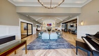 Sutton Landing at Uniondale's clubhouse at 2000 Sutton Court offers a sophisticated, single-floor amenity space designed with upscale finishes and thoughtful detail throughout. The centerpiece is a grand lounge featuring a soaring vaulted ceiling with decorative white beams, rich herringbone-patterned hardwood flooring, and a striking built-in fireplace framed by illuminated bookshelves and a mounted flat-screen television. Plush seating arrangements, elegant table lamps, and a boldly patterned area rug create a warm yet refined gathering atmosphere. An adjoining dining area showcases exposed dark wood ceiling beams, globe pendant lighting, and herringbone hardwood floors, with ample seating for residents to gather and socialize. The fully equipped kitchen features gray cabinetry, quartz countertops, a decorative mosaic tile backsplash, and modern stainless steel appliances. The fitness center is outfitted with cardio equipment, free weights, and large arched windows that invite natural light, complemented by floor-to-ceiling mirrors and teal accent walls. Restrooms are elegantly appointed with hexagonal marble tile flooring, marble wall tile, and iridescent glass mosaic accent tile. Throughout, the clubhouse exudes a cohesive, contemporary coastal aesthetic with a welcoming and polished ambiance.