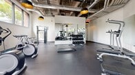 Located at 1 E Pleasant St in Amherst, MA, this fitness center offers a well-equipped, single-floor workout facility designed to support a wide range of exercise routines. The space features an open, industrial-style aesthetic with exposed ductwork and concrete ceilings accented by bold black-and-gold pendant light fixtures, lending the room a modern, urban character.

The floor is finished with durable dark rubber flooring throughout, providing traction and comfort for all types of workouts. Large windows along the perimeter walls flood the space with natural light and offer views of the surrounding greenery, creating an energizing atmosphere.

The cardio zone includes treadmills, elliptical trainers, stationary bikes, and spinning bikes, while the strength training area is equipped with a cable machine, weight benches, adjustable dumbbells, kettlebells, medicine balls, and a dumbbell rack. Full-length wall mirrors amplify the sense of space and allow users to monitor their form. Exercise mats are available for stretching and floor work.

The layout is thoughtfully organized to allow efficient movement between zones without congestion. With its combination of modern equipment, strong natural light, and industrial design, this fitness center provides a functional and motivating environment for residents and users at this Amherst location.