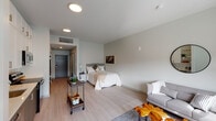 This single-floor unit offers a thoughtfully designed open-concept layout that seamlessly integrates the sleeping, living, and kitchen areas into one cohesive space. Light wood-tone flooring runs throughout, complementing the soft neutral palette of white and grey tones on the walls and ceilings, creating a clean and contemporary aesthetic.

The sleeping area accommodates a full-sized bed and includes a nightstand, with a framed world map artwork serving as a visual anchor on the wall. Adjacent to it, the living area features a grey sofa, a round wall mirror, and a window that draws in natural light and offers views of the surrounding exterior. A unique organic-shaped accent piece adds a touch of character to the lounge space.

The kitchen is well-appointed with two-tone cabinetry — white upper cabinets paired with grey lower cabinets — white subway tile backsplash, light countertops, and a full suite of stainless steel appliances including a range, microwave, dishwasher, and refrigerator.

The bathroom includes a wide vanity with a white countertop, dark lower cabinetry, a large mirror, and a walk-in glass shower, all finished in a clean, modern style. In-unit laundry is also present, as seen in the layout overview.