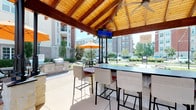 The Grill Area is a well-appointed outdoor entertaining space centered around a striking covered pavilion with a vaulted, exposed-beam wood ceiling that adds warmth and architectural character. The open-air design allows for abundant natural light and a refreshing connection to the surrounding outdoor environment while providing shade and shelter for gatherings. At the heart of the pavilion is a spacious bar-height counter supported by stone bases, surrounded by wicker bar chairs with cushioned seating, creating a comfortable and inviting social hub. A flat-screen television is mounted overhead, making the space equally suited for casual get-togethers and viewing events. Ceiling fans are positioned throughout to ensure airflow during warmer months. Adjacent to the pavilion, a built-in outdoor grill station with ample counter space provides a fully functional cooking setup for outdoor meals. Beyond the covered area, the patio extends outward with additional seating options, including tables shaded by vibrant orange umbrellas. Decorative brick accents border the patio, adding texture and visual interest to the ground-level design. The landscaped surroundings, complete with manicured greenery and planters, frame the space beautifully, while views of the broader courtyard lend an open, community-oriented ambiance to the entire grill area.