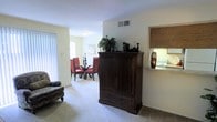 This single-floor residence offers a functional and cohesive layout designed for comfortable everyday living. The open-plan living and dining area forms the heart of the home, creating a seamless flow between spaces. The living area is finished with plush carpeting in soft neutral tones, providing a warm and understated foundation throughout. A large dark wood armoire serves as a prominent focal point in the living space, adding a touch of classic elegance. Vertical blinds on the windows allow for flexible light control, filling the room with soft natural light when open. The adjoining dining area accommodates a round dining table with upholstered chairs in rich red tones, creating a welcoming spot for meals. A framed artwork piece on the wall adds a decorative accent to the dining nook. A pass-through window with a wooden surround connects the living and dining space to the kitchen, enhancing the open feel while maintaining a defined kitchen zone. The kitchen, visible through the pass-through, features white appliances and light cabinetry. Central HVAC venting is visible in the ceiling, ensuring year-round climate comfort. The overall ambiance is calm, tidy, and well-organized, making this a practical and inviting single-floor living space.