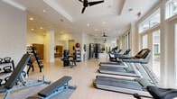 Located at 3500 Fairmount St in Dallas, Texas, this fitness center offers a thoughtfully designed workout environment finished with light hardwood floors, tray ceilings, and recessed lighting. Multiple ceiling fans are distributed throughout, keeping the space comfortable during workouts. Large windows along one wall flood the interior with natural light and provide views of the surrounding outdoor area, contributing to an open, energizing atmosphere. The space is comprehensively equipped for a full range of exercise modalities. A row of treadmills lines one side of the floor, accompanied by stationary bikes and additional cardio equipment. The strength training section features a multi-station cable machine, a leg press, adjustable benches, and a dedicated free weights area with an organized dumbbell rack and kettlebell station. Medicine balls and a ball rack add variety for functional training. Full-length mirrors are strategically placed to expand the sense of space and assist with form. A wall-mounted flat-screen TV is positioned against a textured, patterned accent wall, adding a modern aesthetic touch. A separate alcove area with warm wood cabinetry provides additional storage or amenity space. White columns and crown molding lend an upscale, polished character to the overall design of this well-appointed fitness facility.