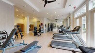 Located at 3500 Fairmount St in Dallas, Texas, this fitness center offers a thoughtfully designed workout environment finished with light hardwood floors, tray ceilings, and recessed lighting. Multiple ceiling fans are distributed throughout, keeping the space comfortable during workouts. Large windows along one wall flood the interior with natural light and provide views of the surrounding outdoor area, contributing to an open, energizing atmosphere. The space is comprehensively equipped for a full range of exercise modalities. A row of treadmills lines one side of the floor, accompanied by stationary bikes and additional cardio equipment. The strength training section features a multi-station cable machine, a leg press, adjustable benches, and a dedicated free weights area with an organized dumbbell rack and kettlebell station. Medicine balls and a ball rack add variety for functional training. Full-length mirrors are strategically placed to expand the sense of space and assist with form. A wall-mounted flat-screen TV is positioned against a textured, patterned accent wall, adding a modern aesthetic touch. A separate alcove area with warm wood cabinetry provides additional storage or amenity space. White columns and crown molding lend an upscale, polished character to the overall design of this well-appointed fitness facility.