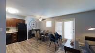 This single-floor residence offers a thoughtfully designed open-concept layout that seamlessly connects the living, dining, and kitchen areas into one cohesive and functional space. The dining area is a standout feature, accommodating a generously sized table with a mix of stylish tan and navy accent chairs, complemented by a bold oversized wall clock that adds a contemporary decorative touch. Natural light pours in through multiple windows fitted with white horizontal blinds, alongside a set of French doors that open to the exterior, enhancing the bright and airy atmosphere. The ceiling fan overhead provides both comfort and a subtle architectural element to the room. Wood-look plank flooring runs throughout, offering warmth and durability underfoot while lending a modern yet welcoming aesthetic. Adjacent to the dining space, the kitchen features dark cabinetry and a full-size black refrigerator, providing a sleek contrast to the lighter tones of the surrounding space. The neutral wall palette and clean lines throughout create a versatile backdrop that balances both style and practicality, making this a comfortable and visually appealing living environment.