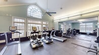 This fitness center is housed in a spacious, single-floor facility distinguished by its vaulted ceilings and classic architectural detailing, including crown molding and a prominent arched window that floods the cardio area with natural light. The walls are finished in a soft teal tone with dark wainscoting below, creating a clean and energizing atmosphere throughout.

The cardio zone is anchored by multiple treadmills and elliptical machines from True Fitness, positioned along a wall of large, multi-pane windows overlooking outdoor greenery. A ceiling fan overhead aids in air circulation, while floor-to-ceiling mirrors along one wall visually expand the space and allow users to monitor their form.

The strength training area is generously equipped with a multi-station cable machine, a full rack of dumbbells, adjustable benches, and various selectorized machines. Durable rubber flooring covers the entire workout floor, providing safety and sound absorption. Built-in shelving along one wall offers convenient storage, and recessed and track lighting ensures the space is well-illuminated throughout. A glass-paned exit door provides easy access, and posted signage keeps the facility organized and user-friendly.