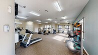 This fitness center provides a generously sized, single-floor workout space designed to accommodate a comprehensive range of exercise needs. The room features a patterned carpet throughout the main exercise area, transitioning to tile flooring near the entry, and is brightened by an abundance of overhead fluorescent lighting that ensures consistent illumination across the entire space. A signature teal accent wall adds a vibrant, energizing touch to the interior, complemented by neutral beige tones on the surrounding walls. Large windows along multiple walls allow natural light to fill the room, contributing to an open and motivating atmosphere. The facility is equipped with a variety of cardio machines, including multiple treadmills and a stationary bike, as well as a cable pulley strength training system and a multi-function weight machine. A dedicated free weights area features a dumbbell rack, medicine balls in multiple weights stored on a vertical rack, and stability balls for functional training. Wall-mounted mirrors on one side of the room enhance the sense of space and allow users to monitor their form. A wall-mounted television is also present for entertainment during workouts. Multiple closet storage areas and a hand sanitizer dispenser reflect thoughtful, user-centered design. The space is well-ventilated with multiple ceiling vents throughout.