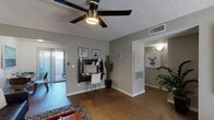 This thoughtfully designed single-floor residence offers a comfortable and functional layout with a cohesive modern aesthetic. The main living area is anchored by rich wood-look flooring, a ceiling fan with integrated lighting, and a sliding glass door that opens to an outdoor patio, filling the space with natural light. A wall-mounted desk nook provides a built-in workspace, adding practical versatility to the room. Adjacent to the living area, the dining space accommodates a round table and chairs beneath warm overhead lighting, with colorful wall art lending a welcoming touch. The kitchen features gray cabinetry, clean white countertops, a double-basin stainless steel sink, and a gas range, offering an efficient and well-organized cooking space. The primary bedroom is carpeted and restful, benefiting from a large window that invites natural light, while the secondary bedroom comfortably fits two beds with its own window and soft carpeting. The full bathroom is neatly appointed with a vanity, wide mirror, and a shower-tub combo. Throughout the residence, neutral gray walls, textured ceilings, and consistent flooring choices create a unified and inviting ambiance suitable for everyday living.