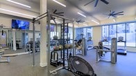 This fitness center is a thoughtfully designed, single-floor facility built to support a wide range of workout routines. The space is outfitted with an impressive array of equipment, including a multi-functional cable and pull-up rig, medicine balls, free weights, and a mini trampoline for functional training. Cardio machines, including treadmills and a stair climber, are positioned near large windows that allow generous natural light to fill the room, creating an energizing and open atmosphere. The flooring features durable rubber matting suited for high-impact activity, while full-length mirrors along the walls enhance the sense of space and allow exercisers to monitor their form. Ceiling fans are mounted throughout to maintain airflow and comfort during workouts, complemented by bright linear lighting that ensures the space is well-lit at all hours. Wall-mounted televisions provide entertainment during cardio sessions. The overall ambiance is clean, modern, and functional, with a layout that offers both open floor space for dynamic movements and dedicated zones for strength and cardio training, making it a versatile and well-rounded fitness environment.