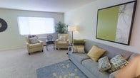 This single-floor living space offers a thoughtfully arranged and inviting environment centered around a comfortable seating area. The room features a grey upholstered sofa adorned with an assortment of decorative pillows in shades of yellow, green, and neutral tones, complemented by two cream-colored armchairs that complete the seating arrangement. A patterned area rug in soft blue and yellow anchors the space, adding a touch of visual interest to the carpeted floor. Natural light filters in through a large window fitted with white horizontal blinds, brightening the room and highlighting the neutral grey-toned walls. A tall floor lamp provides warm ambient lighting, while a bold green-framed artwork on the wall introduces a vibrant accent. Decorative touches, including a potted tropical plant in the corner and a small birdcage ornament on a side table, add personality and warmth to the space. The overall ambiance is calm, clean, and well-coordinated, making this living area an ideal setting for relaxation and everyday living.