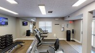Located at 10175 Outrigger Lane in Fishers, Indiana, this fitness center is a single-floor facility designed to support a well-rounded workout experience. The space is organized into distinct workout zones, with neutral gray walls, a combination of carpet and hard flooring, and bright flush-mounted fluorescent lighting throughout that ensures clear visibility across the entire area.

The cardio zone is prominently featured and houses a row of Matrix treadmills and elliptical machines, positioned to face a wall-mounted television and a large mirror that visually expands the space. A free weight section with a dumbbell rack is reflected in the mirrored wall, making the area feel more open. Additional equipment includes stationary bikes, stability balls, medicine balls, and resistance accessories, offering options for both strength and functional training.

Safety and convenience are well-considered throughout the space. A water cooler station, hand sanitizer dispenser, and an AED unit with first aid supplies are mounted on the walls. Clearly posted fitness center policies and multiple exit doors—including a glass-paneled entry—provide easy access and transparency. A window allows natural light to supplement the overhead lighting. The overall environment is clean, functional, and purpose-built for community fitness use.