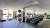 This single-level fitness center is designed to support a comprehensive workout experience in a clean, modern environment. The space is anchored by a row of elliptical trainers and treadmills arranged along mirrored walls, which visually expand the room and allow users to monitor their form during exercise. The flooring consists of durable, cushioned blue-grey tiles that provide comfort and stability underfoot — a practical choice well-suited to high-traffic fitness use.

Large windows along one side of the room invite abundant natural light, creating an open and energizing atmosphere. Recessed ceiling lighting supplements the natural light, ensuring the space remains well-lit throughout. The ceiling features a stepped architectural detail that adds a sense of depth and definition to the room.

A medicine ball rack positioned prominently in the floor space holds multiple exercise balls in varying sizes, catering to functional training and core workouts. Additional cardio and strength equipment is visible toward the rear of the space, indicating a diverse range of training options. The mirrored column at the center of the room serves both a structural and aesthetic purpose, reinforcing the spacious feel of the fitness center overall.