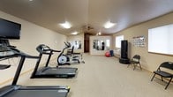 This single-floor fitness and wellness facility offers a comprehensive setup for both exercise and relaxation. The main fitness area is a generously sized room with neutral-toned walls, carpeted flooring, and bright overhead fluorescent lighting complemented by a ceiling fan. The space is outfitted with multiple treadmills, an elliptical trainer, a stationary bike, and a multi-station cable weight machine, along with free-standing strength equipment. A large wall-mounted mirror spans one section of the room, allowing users to monitor their form during workouts. A window provides natural light, and a wall-mounted HVAC unit ensures climate control. Motivational framed artwork and posted instructional guides contribute to the functional atmosphere. Adjoining the main floor is a private tanning room, compact in size and accessed through a wood-framed door. It houses a Royal Sun 32/0 Express tanning bed as its centerpiece. The room includes a wall-mounted coin-operated or card payment unit, a small dark wood storage cabinet with wicker baskets holding sanitizing supplies, a folding wooden chair, and decorative floral wall art, creating a tidy and practical personal care space.