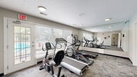 Located at 8016 E Louisville Dr in Lawrence, Indiana, this dedicated exercise room offers a practical and well-organized fitness space designed to support a range of cardio-focused workouts. The room is outfitted with multiple pieces of equipment, including treadmills, elliptical trainers, and a stationary bike, providing users with varied options for cardiovascular exercise. Large windows fitted with horizontal blinds line one wall, allowing an abundance of natural light to fill the space while offering views of the outdoor surroundings, including what appears to be a pool area. A full-length wall mirror spans one side of the room, visually expanding the space and enhancing the gym-like atmosphere. The flooring consists of durable, patterned carpet tiles in a neutral gray tone, well-suited for a high-traffic fitness environment. Recessed and flush-mount ceiling lights provide consistent, even illumination throughout. The textured ceiling and clean, light-painted walls contribute to a bright and functional aesthetic. Two entry doors provide convenient access, with one featuring a windowed panel for added openness. The overall design is straightforward and purpose-driven, making this fitness room a practical amenity for everyday use.