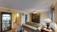 Located at 880 Glendale Dr in Cary, NC, this two-story home offers a comfortable and well-appointed layout designed for everyday living. The main living area serves as the heart of the home, featuring classic crown molding, a textured ceiling, and a ceiling fan that adds both function and character. Warm, neutral wall tones with a two-tone treatment — defined by a chair rail — lend the space a timeless, traditional feel. Hardwood flooring anchors the living area, adding warmth and durability underfoot. The living room flows naturally toward the kitchen, which is visible through a connecting doorway and equipped with appliances, making it convenient for daily use. A hallway off the main living space provides access to additional rooms, including what appears to be a closeted space, suggesting well-organized storage throughout the home. The open sightlines between rooms promote a sense of connectivity and ease of movement across the floor plan. With its classic architectural details, warm color palette, and practical layout spread across two floors, this Cary home presents a welcoming and functional environment suited to a variety of living arrangements.