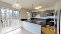 This single-floor residence offers a clean, modern design with a thoughtful layout that balances open communal spaces and private rooms. The kitchen is a standout feature, showcasing sleek gray shaker-style cabinetry, dark granite countertops, and a full suite of stainless steel appliances including a refrigerator, range, and built-in microwave. A peninsula with additional granite counter space extends into the living area, creating a natural divide while maintaining an open, connected feel. Pendant lighting fixtures and a flush-mount ceiling light illuminate the kitchen, which benefits from natural light streaming through a sliding glass door that opens to an outdoor area. The living and dining areas are finished with light hardwood-style flooring and bright white walls, creating a spacious, airy ambiance. A staircase with a wood handrail and black metal balusters adds architectural interest. The bedrooms feature neutral-toned carpet and windows that invite natural light. The bathroom is finished in clean white tones with a full soaking tub and shower combination, tile-style flooring, and a window for ventilation and light. Throughout the space, the consistent use of neutral finishes and natural light creates a welcoming and functional environment.