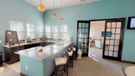 This single-floor community space is thoughtfully designed to offer a warm, inviting atmosphere with a blend of classic and contemporary elements. The main lounge area features soaring ceilings, a multi-arm chandelier, and large double entry doors that flood the space with natural light. Neutral-toned sofas, accent chairs, and a mix of area rugs create comfortable conversation areas throughout.

A standout feature is the floor-to-ceiling stacked stone fireplace, complete with a rustic wood mantle and decorative accessories, anchoring a cozy seating nook. French doors adjacent to the fireplace open directly to an outdoor patio, enhancing the connection between interior and exterior spaces.

The kitchen and bar area is finished in a vibrant aqua blue, offering a refreshing pop of color. Dark cabinetry with granite countertops lines the walls beneath large plantation-shuttered windows, while pendant lights with warm amber shades hang above a spacious bar counter with seating for several guests—ideal for entertaining or casual gatherings.

Dark-trimmed French doors with glass panes connect the various zones of the space, maintaining visual openness while defining individual areas. Throughout, the design palette of soft whites, aqua blues, and warm wood tones creates a cohesive, polished ambiance suited for both social events and everyday use.