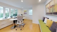 This well-appointed single-floor office space is thoughtfully designed to support both focused work and collaboration. The main workspace is anchored by an L-shaped built-in desk running along the walls beneath a series of large windows, providing two fully equipped workstations with ample surface area for monitors, phones, and office equipment. Natural light pours in generously through multiple windows, including a wide bay-style window that frames lush, green outdoor views, creating a bright and energizing atmosphere throughout the day. Recessed ceiling lighting ensures consistent illumination at all hours. An oval white pedestal table with chrome-accented ergonomic chairs occupies the center of the room, offering a flexible space for meetings or collaborative tasks. Climate comfort is addressed by a wall-mounted mini-split air conditioning unit. A standout feature is the built-in bench along one wall, upholstered in a vibrant green fabric with stylish throw pillows, topped by a large framed abstract artwork in warm yellow and earth tones — adding a welcoming, creative touch to the space. Light hardwood flooring runs throughout, complementing the clean white walls and cabinetry. A private door provides direct access to the outdoors, enhancing the sense of openness and convenience.