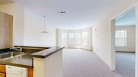 This 600-square-foot, one-bedroom, one-bathroom apartment at Union Place in Franklin, MA offers a practical and comfortable single-floor layout. The living room is a standout feature, boasting a curved bay window configuration that floods the space with natural light and provides pleasant outdoor views of surrounding greenery. Neutral beige walls and plush wall-to-wall carpeting throughout the living and bedroom areas create a warm, cohesive ambiance. The kitchen features warm honey-toned wood cabinetry, a dark countertop, white appliances including a full-size refrigerator and oven, and light tile flooring, providing ample storage and functionality. The 170-square-foot bedroom includes a large closet with a built-in rod and shelving system, carpeted flooring, and a window offering natural light. The bathroom is finished with tan ceramic tile flooring, a vanity with cabinet storage, and accessibility-friendly grab bars. A notable convenience is the in-unit stacked washer and dryer located adjacent to the bathroom. The apartment's clean, neutral palette and functional layout make it an efficient and comfortable living space.