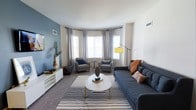 This 2-bedroom, 2-bathroom single-floor residence offers a thoughtfully designed living space with a modern aesthetic. The living room serves as the focal point, showcasing a curved bay window arrangement that allows abundant natural light to fill the room, framed by elegant grey curtains. A bold slate-blue accent wall adds depth and character, complementing the neutral tones throughout. The space accommodates a generous navy sectional sofa paired with coordinating grey accent chairs, centered around a round white coffee table atop a classic black-and-white striped area rug. A sleek white media console lines one wall, providing functional storage while maintaining the clean, contemporary feel. A statement yellow arc floor lamp introduces a warm pop of color, enhancing the inviting ambiance. The carpeted flooring contributes to the comfortable and cozy atmosphere of the living area. The overall design balances modern furnishings with practical layout choices, creating a welcoming environment suited for both relaxing and entertaining. The unit's single-floor layout ensures easy accessibility and a cohesive flow throughout the living spaces.