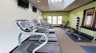 This fitness center is designed to support a well-rounded workout experience within a clean, purpose-built environment. The space features high ceilings with a raised soffit detail and recessed lighting, contributing to an open and airy atmosphere. Neutral-toned carpeting lines the floor, while accent walls painted in a fresh sage green add a sense of energy and vitality to the room. Natural light filters in through a row of windows along one wall, brightening the interior and creating a welcoming workout environment. Wall-mounted flat-screen televisions are positioned for easy viewing from the cardio equipment.

The cardio section is prominently featured, housing a row of Star Trac treadmills and elliptical trainers arranged in an orderly line. Stationary and recumbent bikes are positioned toward the windowed wall, offering a comfortable option for lower-impact exercise. Functional training accessories, including a balance dome and exercise mat, are available for stretching and core work. The layout is thoughtfully arranged to maximize the use of the available floor space while maintaining clear pathways between equipment. Overall, the fitness center presents a practical and well-maintained facility suited for a variety of cardio and fitness training needs.