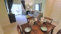 This single-floor residence showcases an open-concept layout that seamlessly connects the dining and living areas, creating a cohesive and functional living space. The dining area is anchored by a rich, dark wood table surrounded by matching chairs, elegantly set with green ceramic bowls and stemware, lending a polished, welcoming feel.

The living area flows naturally from the dining space, furnished with a neutral gray sofa accented by patterned blue and green throw pillows. A coordinating upholstered armchair and ottoman complete the seating arrangement, while a decorative framed mirror adds visual depth to the wall above. A table lamp provides warm ambient lighting to complement the natural light in the room.

Large sliding glass doors along one wall allow abundant natural light to fill the space and provide direct access to a private outdoor patio, where additional seating is visible. Light-toned carpeting throughout contributes to the bright, airy ambiance. The neutral color palette of soft greens and warm beiges, paired with carefully chosen furnishings, creates an inviting and well-balanced atmosphere throughout the home.