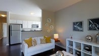 Located at 5730 Silver Falls St in Dublin, Ohio, this single-floor residence offers a thoughtfully designed living space with a clean, contemporary aesthetic. The living room serves as the heart of the home, finished in cool gray tones that lend a calm, modern atmosphere. Dark hardwood-style flooring grounds the space and contrasts elegantly with the light furnishings, including a white sofa adorned with mustard yellow and patterned accent pillows that introduce warmth and personality. A striped area rug further defines the seating area, while a white open-cube shelving unit displays decorative accents and provides practical storage. Metallic spherical décor pieces and framed wall art add visual interest throughout. Ambient lighting from a tall floor lamp and a table lamp creates a warm, inviting glow. Decorative circular mirrors arranged on one wall add depth and a sculptural quality to the room. An open pass-through connects the living area to the kitchen, which features white cabinetry, dark countertops, and stainless steel appliances, including a built-in microwave. The seamless flow between the living and kitchen areas makes the space feel open and connected, ideal for both everyday living and entertaining.