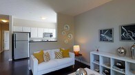 Located at 5730 Silver Falls St in Dublin, Ohio, this single-floor residence offers a thoughtfully designed living space with a clean, contemporary aesthetic. The living room serves as the heart of the home, finished in cool gray tones that lend a calm, modern atmosphere. Dark hardwood-style flooring grounds the space and contrasts elegantly with the light furnishings, including a white sofa adorned with mustard yellow and patterned accent pillows that introduce warmth and personality. A striped area rug further defines the seating area, while a white open-cube shelving unit displays decorative accents and provides practical storage. Metallic spherical décor pieces and framed wall art add visual interest throughout. Ambient lighting from a tall floor lamp and a table lamp creates a warm, inviting glow. Decorative circular mirrors arranged on one wall add depth and a sculptural quality to the room. An open pass-through connects the living area to the kitchen, which features white cabinetry, dark countertops, and stainless steel appliances, including a built-in microwave. The seamless flow between the living and kitchen areas makes the space feel open and connected, ideal for both everyday living and entertaining.