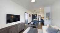 This stylish single-floor residence showcases a thoughtfully designed open-concept layout that connects the living area and kitchen in one cohesive, functional space. Dark hardwood floors run throughout, grounding the interior with warmth and sophistication. The living area is bright and inviting, featuring recessed lighting and a ceiling-mounted light fixture that together provide ample illumination. A wall-mounted flat-screen TV sits atop a rustic, dark wood media console, adding a touch of contrast against the clean white walls. A patterned area rug defines the seating zone, complementing the neutral tones of the sofa. The kitchen is a standout feature, boasting white upper cabinetry, dark lower cabinetry, and a central island with bar seating. A vessel sink rests atop the island, adding a distinctive design element. Stainless steel appliances, including a full-size refrigerator, complete the modern culinary space. Pendant lighting above the island adds both ambiance and function. Interior doors with dark trim frames provide visual continuity throughout the space. The overall aesthetic is contemporary and cohesive, blending dark and light tones for a balanced, urban feel.