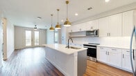 This well-designed two-bedroom, two-bathroom residence in Austin, TX offers a thoughtfully laid-out single-floor plan with modern finishes throughout. The open-concept kitchen and living area serves as the heart of the home, anchored by a large center island with a white quartz countertop, an undermount sink, and striking gold pendant lights that add a warm, contemporary touch. White shaker-style cabinetry, a classic subway tile backsplash, and stainless steel appliances — including a range and built-in microwave — complete the kitchen. Rich wood-look flooring flows seamlessly throughout the entire space, complementing the clean, neutral color palette. The living area is bright and airy, benefiting from natural light that pours in through glass French doors leading to an outdoor balcony. Both bedrooms are generously sized with crisp white walls and the same warm flooring, offering a calm and versatile canvas. The primary suite includes a dedicated walk-in closet outfitted with chrome hanging rods, as well as an en-suite bathroom featuring a glass-enclosed shower with penny tile detailing and matte black fixtures. A hallway connects all rooms efficiently, and two closets provide ample storage throughout the home.