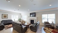 This single-floor space is designed as a comfortable and functional communal lounge area. The room features a classic white-mantled fireplace with an elegant stone surround, serving as a natural focal point. A large wall-mounted flat-screen television is positioned above the fireplace, making it ideal for group viewing. The space is furnished with dark leather sofas and chairs arranged around a rich wood coffee table, creating an inviting seating arrangement suited for conversation or relaxation. Additional accent chairs in neutral tones are positioned near the windows, offering quieter spots to sit. The flooring features a speckled terrazzo-style finish that adds texture and durability to the space. Recessed lighting in the white ceiling provides even illumination throughout, while multiple large windows fitted with white horizontal blinds allow generous natural light to fill the room. The neutral color palette of whites and grays throughout the walls and furnishings contributes to a clean, contemporary aesthetic. A decorative plant near the window adds a touch of greenery. Overall, the space strikes a balance between comfort and practicality, well-suited for shared use.