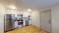 This single-floor unit offers a clean, modern aesthetic throughout its thoughtfully designed layout. The kitchen is a standout feature, equipped with a full suite of stainless steel appliances including a refrigerator, electric range, and dishwasher. Crisp white cabinetry provides ample storage, while granite-style countertops and a geometric-patterned tile backsplash add a contemporary touch. Light wood-tone flooring runs throughout the kitchen and living areas, lending warmth and visual continuity to the space.

The living area is open and airy, benefiting from the kitchen's flush ceiling lights that keep the combined space bright and inviting. A dedicated entry door provides a clear separation from the main living zone.

The bedroom is a calm, neutral retreat featuring light wood-tone flooring, a large window dressed with horizontal blinds that allows natural light to filter in, and a wall-mounted air conditioning unit alongside baseboard heating for year-round comfort. A ceiling-mounted smoke detector adds a practical safety element.

The bathroom is finished with white tile surrounding a full bathtub and shower combination, accented by a vertical strip of elongated hexagonal brown decorative tiles, creating an elegant focal point. The overall design of the unit balances functionality with modern, clean-lined finishes throughout.