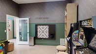 This single-floor amenity space is a thoughtfully designed entertainment hub that blends retro gaming culture with modern interior design. The main game room is immediately eye-catching, featuring bold two-tone walls with dark, floral-patterned wallpaper on the upper half and rich forest-green wainscoting below — a combination that sets a playful yet sophisticated tone throughout. A gold-lettered "Work Hard. Play Hard." motto is mounted prominently on the feature wall, reinforcing the space's energetic character.

The game room is outfitted with a curated collection of classic arcade cabinets, including Atari 50, Pac-Man, and Pong machines, accompanied by round upholstered stools for casual seating. A large wall-mounted Scrabble board adds a social, analog counterpart to the digital games. A natural wood open bookshelf provides storage for board games and accessories.

Light herringbone-pattern flooring runs throughout, adding warmth and visual texture underfoot. A pass-through window connects to an adjacent space, while a set of double barn doors with a rustic natural wood finish offer a charming dividing element. Recessed lighting illuminates the space cleanly from above. A glass-paneled privacy pod is also accessible, rounding out this well-appointed, multi-use recreational area.