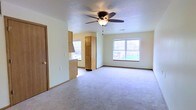 This single-story space features an open-concept layout that seamlessly connects the living area to the kitchen. The living room is spacious and carpeted, offering a comfortable, understated aesthetic with neutral walls and a textured ceiling. A ceiling fan with an integrated light fixture is centrally mounted, providing both air circulation and ambient lighting.

Two windows allow natural light to fill the room, creating a bright and welcoming atmosphere. Views through the windows suggest a residential outdoor setting. The room also includes a dedicated door along one wall, likely leading to additional areas or an exterior entry.

The kitchen is partially visible through a pass-through counter, which promotes an open flow between the cooking and living spaces. Light wood cabinetry, including a tall pantry-style cabinet, provides ample storage. The kitchen benefits from its own window, ensuring good natural light for meal preparation.

Overall, the space presents a functional and practical layout with clean lines, warm wood tones, and neutral finishes that create a welcoming, move-in-ready environment suited to everyday living.