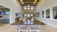 This thoughtfully designed single-story community space blends coastal charm with functional amenities across a generous, open floor plan. The heart of the facility is a dramatic great room anchored by soaring ceilings with exposed wooden beams, clerestory windows, and a striking dual-blade ceiling fan that draws the eye upward. Soft sage-green walls, light wood-look tile flooring, and carefully curated furnishings create a welcoming, resort-like atmosphere throughout.

The main gathering area flows seamlessly into a sun-drenched dining nook enclosed by a curved bay of floor-to-ceiling windows dressed in patterned drapes, offering views of the surrounding outdoor spaces. A large high-top table with whitewashed legs and a dark metal top serves as a central social hub nearby. Adjacent seating areas feature plush neutral sofas, armchairs, and patterned area rugs arranged for comfortable conversation.

A dedicated lounge room with a bold teal ceiling, recessed lighting, and multiple wall-mounted televisions provides a cozy media retreat. The refreshment station includes teal cabinetry, countertop beverage dispensers, and additional mounted screens. A game room with a billiard table, bright entry foyer with glass panel doors, and interior office windows round out this well-appointed facility.