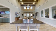 This thoughtfully designed single-story community space blends coastal charm with functional amenities across a generous, open floor plan. The heart of the facility is a dramatic great room anchored by soaring ceilings with exposed wooden beams, clerestory windows, and a striking dual-blade ceiling fan that draws the eye upward. Soft sage-green walls, light wood-look tile flooring, and carefully curated furnishings create a welcoming, resort-like atmosphere throughout.

The main gathering area flows seamlessly into a sun-drenched dining nook enclosed by a curved bay of floor-to-ceiling windows dressed in patterned drapes, offering views of the surrounding outdoor spaces. A large high-top table with whitewashed legs and a dark metal top serves as a central social hub nearby. Adjacent seating areas feature plush neutral sofas, armchairs, and patterned area rugs arranged for comfortable conversation.

A dedicated lounge room with a bold teal ceiling, recessed lighting, and multiple wall-mounted televisions provides a cozy media retreat. The refreshment station includes teal cabinetry, countertop beverage dispensers, and additional mounted screens. A game room with a billiard table, bright entry foyer with glass panel doors, and interior office windows round out this well-appointed facility.
