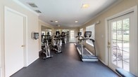 This fitness center offers a comprehensive workout environment housed within a single-floor layout designed to accommodate a wide range of exercise routines. The space features durable dark rubber flooring throughout, providing both safety and functionality for high-traffic athletic use. Crown molding and neutral warm-toned walls lend a polished, well-maintained appearance to the room.

The cardio section is equipped with multiple treadmills and elliptical machines from True Fitness, as well as a row of spin bikes, giving users ample options for sustained cardiovascular training. A multi-station cable and strength machine anchors the resistance training area, complemented by a weight bench and additional free weight equipment, offering a full-body strength training setup.

Large wall-mounted mirrors run along one side of the room, visually expanding the space and allowing users to monitor their form during workouts. Wall-mounted flat-screen televisions are positioned for easy viewing from multiple exercise stations, adding an entertainment element to training sessions. Natural light filters in through several windows fitted with horizontal blinds, and recessed ceiling fixtures provide consistent artificial lighting. A sanitizing station is conveniently mounted near the entrance. A glass-paneled door at one end of the space offers access to the exterior, suggesting proximity to an outdoor amenity area.