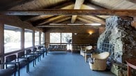 This single-floor rustic retreat exudes warm, lodge-style character defined by its striking architectural details and natural materials throughout. The soaring vaulted ceiling is supported by prominent exposed timber beams, lending a sense of grandeur and craftsmanship to the interior. The walls are clad in rich, horizontal wood planking that reinforces the cabin aesthetic and creates a cohesive, earthy atmosphere.

A commanding stacked-stone fireplace anchors one end of the main gathering area, serving as a natural focal point and adding to the cozy ambiance of the space. Opposite the fireplace, a generous arrangement of large windows lines the walls, flooding the room with natural light and offering immersive views of the surrounding wooded landscape. The connection to nature is a defining feature of this space.

The main area is furnished with a versatile mix of seating, including sofas, armchairs, and wicker chairs, arranged to encourage both communal gatherings and relaxed conversation. Additional row-style seating along one side suggests the room's adaptability for organized events or meetings. Blue-grey carpeting grounds the space and softens the otherwise rugged textures of wood and stone. Wall-mounted sconces provide warm supplemental lighting, enhancing the inviting character of this distinctive, nature-immersed retreat.