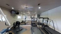 This single-level fitness room offers a comprehensive workout environment designed to accommodate a variety of exercise routines. The space is lined with large floor-to-ceiling mirrors along one wall, which visually expand the room and allow users to monitor their form during exercise. Recessed lighting throughout the ceiling, supplemented by a centrally mounted ceiling fan, ensures the room remains well-lit and comfortable during workouts. The flooring combines protective rubber mats in key equipment zones with standard carpeting in surrounding areas, providing both safety and practicality. Cardio equipment, including multiple treadmills, is positioned prominently within the space, while strength training equipment and free weights, including dumbbells and a weight bench, are arranged along the perimeter. A wall-mounted television adds an entertainment option for users during extended sessions. The layout is organized to allow clear circulation between equipment, making the space functional and accessible. Overall, the fitness room presents a clean, utilitarian design focused on supporting a broad range of fitness activities.
