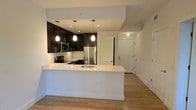 This single-floor space showcases a thoughtfully designed open-concept layout that seamlessly connects the kitchen to the living area. The kitchen serves as a focal point, featuring dark espresso cabinetry that provides a striking contrast against crisp white countertops. A generously sized peninsula with an undermount sink offers both additional prep space and a natural gathering spot. Stainless steel appliances, including a refrigerator, microwave, and range, add a polished, contemporary feel. Three pendant lights hang above the peninsula, providing focused task lighting while enhancing the kitchen's visual appeal. Recessed lighting throughout further brightens the space. Light-toned hardwood flooring runs continuously through the kitchen and into the adjacent living area, contributing to a cohesive and airy atmosphere. The open sightlines create a sense of spaciousness, making the layout feel connected and functional. White walls and trim throughout maintain a clean, neutral palette that complements the modern design elements. The space also features multiple doorways, offering practical access to additional rooms and storage areas.