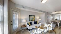 Aspenwood Apartments presents a thoughtfully designed single-floor residence that combines contemporary style with comfortable living. The open living area serves as the heart of the space, featuring light wood-toned flooring that runs seamlessly throughout and neutral gray walls that create a calm, sophisticated backdrop. Large windows dressed with horizontal blinds allow generous amounts of natural light to fill the room, enhancing the bright and airy atmosphere. The living area comfortably accommodates a seating arrangement centered around a geometric-patterned area rug, complemented by natural wood accent tables and warm ambient lighting from stylish table lamps. A bold abstract artwork anchors the main wall, adding a pop of color and artistic character to the space. The smooth white ceilings with clean lines and recessed HVAC vents contribute to the polished, well-maintained aesthetic. An open passage leads to additional areas of the unit, suggesting a flowing, connected floor plan. Overall, the space exudes a modern yet inviting ambiance, ideal for those seeking a refined and comfortable living environment at Aspenwood Apartments.
