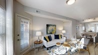 Aspenwood Apartments presents a thoughtfully designed single-floor residence that combines contemporary style with comfortable living. The open living area serves as the heart of the space, featuring light wood-toned flooring that runs seamlessly throughout and neutral gray walls that create a calm, sophisticated backdrop. Large windows dressed with horizontal blinds allow generous amounts of natural light to fill the room, enhancing the bright and airy atmosphere. The living area comfortably accommodates a seating arrangement centered around a geometric-patterned area rug, complemented by natural wood accent tables and warm ambient lighting from stylish table lamps. A bold abstract artwork anchors the main wall, adding a pop of color and artistic character to the space. The smooth white ceilings with clean lines and recessed HVAC vents contribute to the polished, well-maintained aesthetic. An open passage leads to additional areas of the unit, suggesting a flowing, connected floor plan. Overall, the space exudes a modern yet inviting ambiance, ideal for those seeking a refined and comfortable living environment at Aspenwood Apartments.