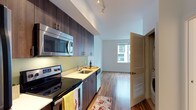 This well-appointed single-floor residence offers a thoughtfully designed layout that balances style and functionality. The kitchen serves as the heart of the space, showcasing dark espresso-toned wood cabinetry that extends to the ceiling, paired with stainless steel appliances including an electric range, built-in microwave, and dishwasher. Light countertops provide a striking contrast to the rich cabinetry, while track lighting overhead ensures the workspace is well-illuminated. Warm hardwood flooring flows seamlessly throughout, connecting the kitchen to the open living area beyond. The living space benefits from natural light pouring in through a large window that frames views of the surrounding cityscape, creating a bright and airy atmosphere. A louvered door leads to an in-unit laundry area, adding a layer of convenience to everyday living. The clean white walls and modern finishes throughout lend the space a crisp, contemporary feel. The open-plan arrangement between the kitchen and living area encourages an easy flow and makes the most of the available footprint, resulting in a cohesive and inviting living environment.