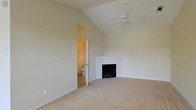 This single-floor space is defined by its dramatic vaulted ceilings, which lend an open, airy quality to the main living area. A ceiling fan is centrally mounted at the peak, providing both circulation and a practical accent to the soaring roofline. The room is finished with soft, neutral-toned carpet and warm beige walls, creating a calm and cohesive atmosphere throughout.

A corner fireplace with a clean white surround and dark firebox serves as a natural focal point, adding warmth and character to the space. The minimalist design of the fireplace integrates seamlessly with the understated aesthetic of the room. An adjoining doorway visible from the main area leads to a bathroom, indicating a connected layout that adds convenience and functionality.

The overall palette of the space is light and understated, relying on architectural features such as the vaulted ceiling and fireplace rather than ornamental detail to create visual interest. The absence of clutter and the open floor plan allow the spatial volume to take center stage, making the area feel generously proportioned and versatile for a variety of uses.