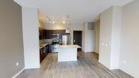 This single-floor residence showcases a thoughtfully designed open-concept layout that seamlessly integrates the kitchen and living areas. The kitchen is a standout feature, equipped with stainless steel appliances including a refrigerator, range, and microwave. Rich dark-toned cabinetry provides ample storage while creating a striking visual contrast against the light granite countertops. A central kitchen island with a pendant light overhead serves as both a functional workspace and a natural gathering point, enhancing the open flow of the space.

Track lighting along the kitchen ceiling ensures excellent task illumination, complementing the warm, neutral tones of the hardwood-style flooring that extends throughout the unit. The walls are finished in a clean, light tone that amplifies the sense of openness and brightness. The living area offers generous space adjacent to the kitchen, providing flexibility for various furniture arrangements. The entryway, visible from the main living area, includes a utility closet, adding practical storage. Overall, the space presents a clean, contemporary aesthetic with a cohesive and functional design.