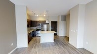 This single-floor residence showcases a thoughtfully designed open-concept layout that seamlessly integrates the kitchen and living areas. The kitchen is a standout feature, equipped with stainless steel appliances including a refrigerator, range, and microwave. Rich dark-toned cabinetry provides ample storage while creating a striking visual contrast against the light granite countertops. A central kitchen island with a pendant light overhead serves as both a functional workspace and a natural gathering point, enhancing the open flow of the space.

Track lighting along the kitchen ceiling ensures excellent task illumination, complementing the warm, neutral tones of the hardwood-style flooring that extends throughout the unit. The walls are finished in a clean, light tone that amplifies the sense of openness and brightness. The living area offers generous space adjacent to the kitchen, providing flexibility for various furniture arrangements. The entryway, visible from the main living area, includes a utility closet, adding practical storage. Overall, the space presents a clean, contemporary aesthetic with a cohesive and functional design.