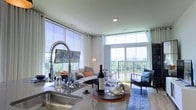 This single-floor space offers an open-concept layout that seamlessly connects the kitchen and living areas. The kitchen is equipped with a sleek countertop, an undermount sink, and a striking industrial-style coil spring faucet, combining functionality with contemporary design. The living area is generously sized, anchored by a plush sofa accented with colorful throw pillows and complemented by a patterned area rug that adds texture and warmth to the space. Large floor-to-ceiling windows wrap around the living area, flooding the interior with abundant natural light and framing sweeping outdoor views of the surrounding landscape. Sheer white curtains line the windows, allowing light to filter softly throughout the space. A glass sliding door provides direct access to a private balcony, extending the living space outdoors and offering an additional seating area. Recessed lighting and smooth, light-toned ceilings contribute to the airy, modern ambiance. The overall aesthetic is clean and contemporary, with a neutral color palette punctuated by curated decorative accents, creating a welcoming and stylish environment.