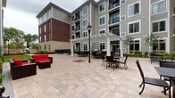 Located at 59 2nd Ave in Raritan, NJ, this outdoor courtyard is a well-appointed communal space nestled within a multi-story residential building. The courtyard is defined by a generously sized, multi-tone stone paver surface that extends across two interconnected zones, providing ample room for both relaxed gatherings and more structured outdoor dining.

A standout feature is the white pergola structure, which anchors one end of the courtyard and shelters a dedicated seating and dining area furnished with wrought-iron tables and chairs. A classic black lamppost adds a charming, pedestrian-friendly aesthetic to the space. On the opposite end, a quieter patio area is framed by fresh landscaping, featuring young trees, ornamental grasses, ferns, and mulched garden beds that introduce natural texture and greenery throughout.

Wooden benches are thoughtfully placed along the perimeter, offering comfortable spots to rest and enjoy the surroundings. Large floor-to-ceiling windows along the building façade provide a visual connection to an interior fitness center, which is visible and directly accessible from the courtyard via glass doors. The multi-story building exterior, clad in neutral gray siding and brick accents, encloses the space on multiple sides, creating a sheltered and intimate atmosphere while still remaining open to the sky above.