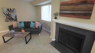 This single-story space offers a well-appointed living area designed for comfort and functionality. The room centers around a classically styled fireplace framed by a crisp white mantel and dark granite surround, providing both a visual focal point and a practical amenity. Above the mantel, the wall offers display space, currently accented with decorative greenery and artwork. Soft, neutral-toned carpeting runs throughout the living area, contributing to a warm and inviting atmosphere. A large window fitted with horizontal blinds allows abundant natural light to fill the space while maintaining privacy. The walls are finished in a warm beige tone, complementing the neutral palette of the room. The layout accommodates comfortable seating and a coffee table, demonstrating efficient use of the available floor space. Overall, the space presents a straightforward, functional design with classic residential features, including the standout fireplace, that combine to create a cozy and livable environment suitable for everyday use.