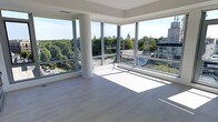 This contemporary space makes an immediate impression with its expansive floor-to-ceiling wraparound glass windows, which flood the interior with abundant natural light and frame stunning panoramic views of the surrounding cityscape and tree canopy. The open layout is characterized by clean, minimalist architecture, featuring smooth white walls, crisp tray ceiling detailing, and a sleek cylindrical structural column that adds an elegant architectural accent without interrupting the sense of openness. Light-toned hardwood flooring runs throughout, complementing the bright, airy atmosphere and reflecting the generous sunlight that streams in throughout the day. The elevated position of the space offers elevated sightlines over neighboring rooftops, verdant treetops, and a dynamic urban horizon, creating a seamless connection between the interior and the world outside. The overall ambiance is one of modern sophistication — uncluttered, luminous, and thoughtfully designed to maximize both space and light. Whether used as a residential or commercial setting, this space offers a rare combination of open-plan flexibility, architectural refinement, and an inspiring outlook that sets it apart.